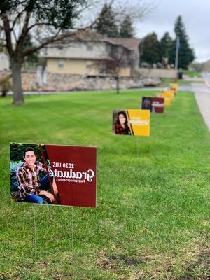 Rectangular signs with images of graduating seniors are arranged in a row across green lawn