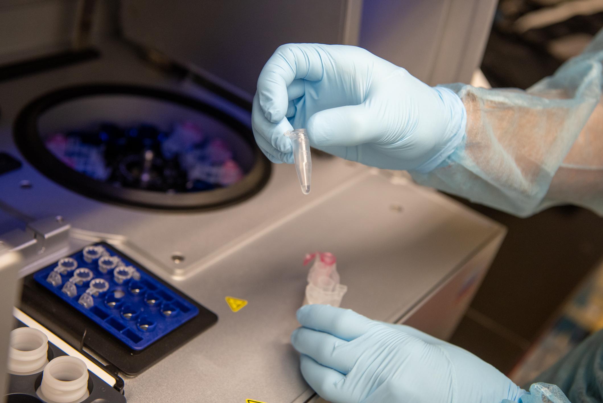 Hand holding a microcentrifuge tube next to the microcentrifuge equipment. There is a tray for the tubes next to the equipment as well.