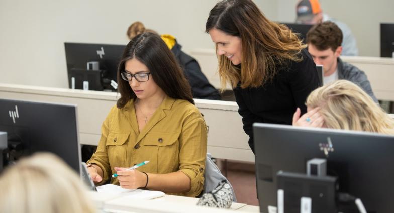 A student sits in front of a computer monitor as a teacher looks over their shoulder.